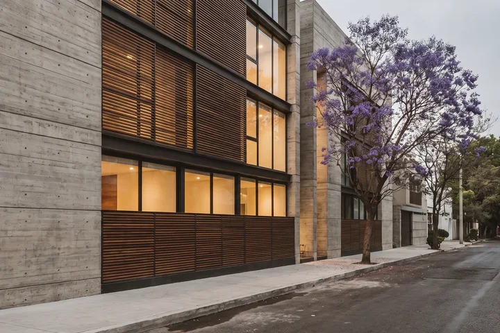 Edificio de concreto aparente y celosías de madera en una calle arbolada con jacarandas